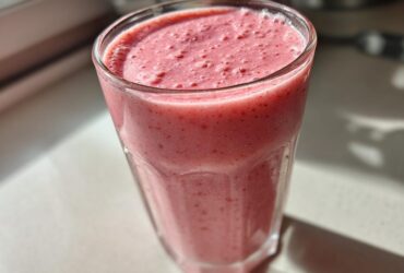 Close-up of a tall glass filled with a thick, pink Keto Strawberry Cheesecake Smoothie, catching bright sunlight.