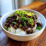 A white bowl filled with white rice topped generously with saucy Korean ground beef bulgogi and garnished with sliced green onions and sesame seeds.