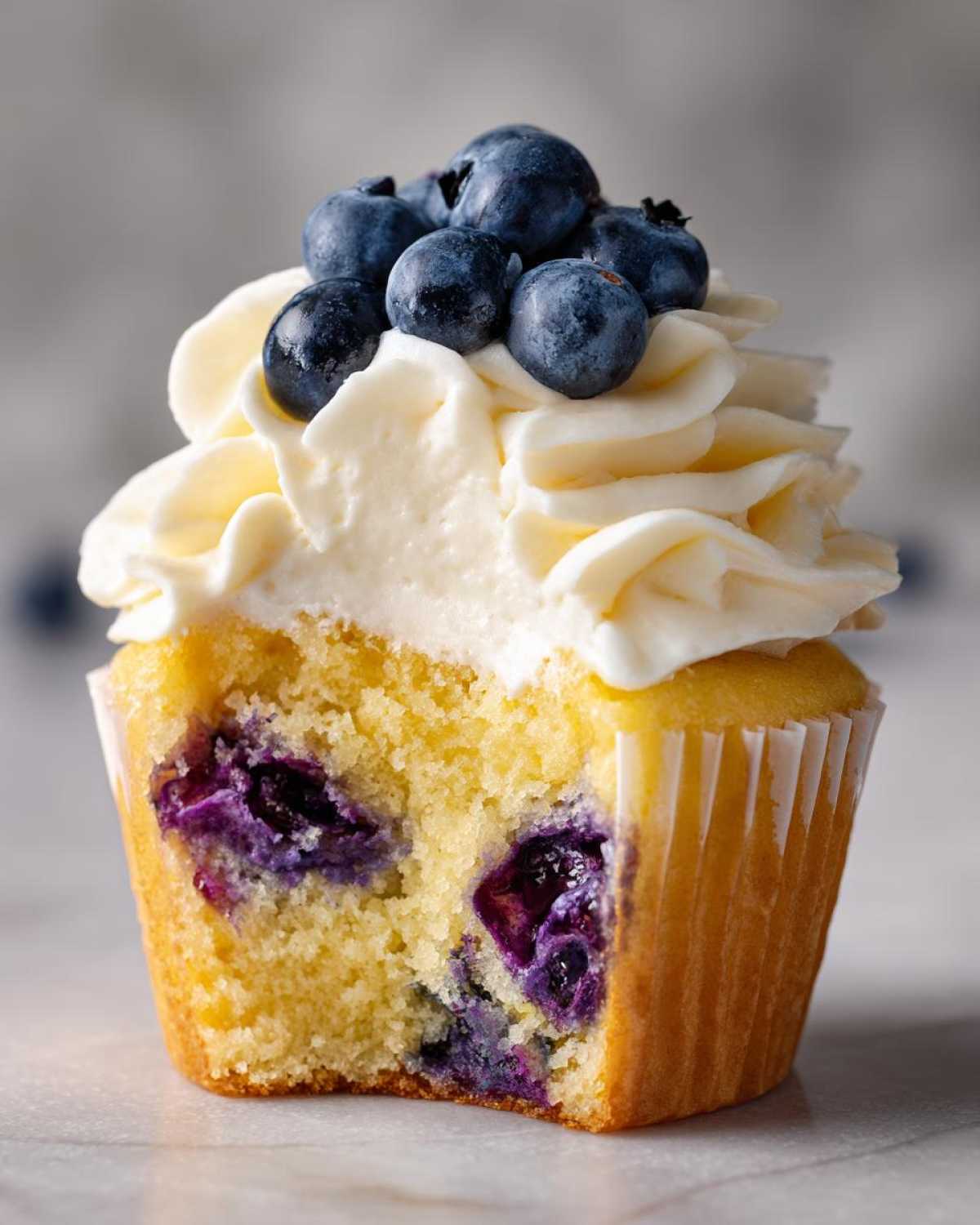 Close-up of a Lemon Blueberry Cupcake with Cream Cheese Frosting, showing a bite taken out to reveal moist cake and burst blueberries.