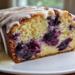 Close-up of a moist slice of Lemon Blueberry Yogurt Loaf, showing blueberries baked inside and topped with a white lemon glaze.