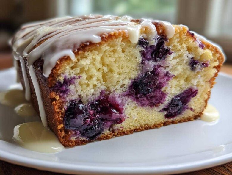Close-up of a moist slice of Lemon Blueberry Yogurt Loaf, showing blueberries baked inside and topped with a white lemon glaze.