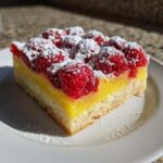 A square slice of Lemon Raspberry Bars featuring a thick lemon layer, fresh raspberries, and powdered sugar dusting.