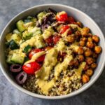 Close-up of a Mediterranean Quinoa Bowl topped with roasted chickpeas, tomatoes, cucumbers, olives, and Lemon Tahini Sauce.
