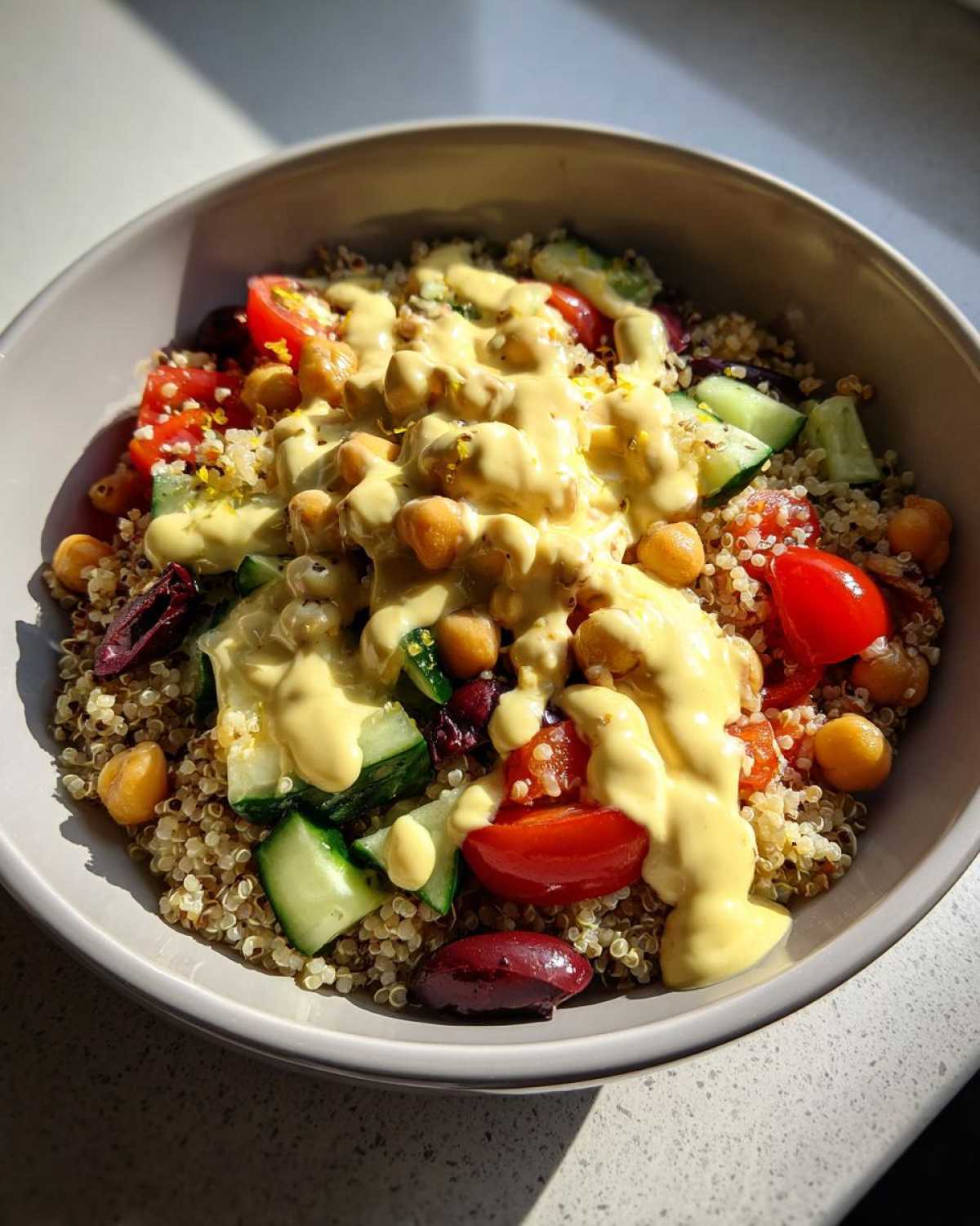 Close-up of a Mediterranean Quinoa Bowl topped generously with creamy lemon tahini sauce, chickpeas, tomatoes, and cucumbers.