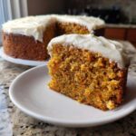 A close-up slice of moist carrot cake with cream cheese frosting on a white plate, with the rest of the cake blurred in the background.