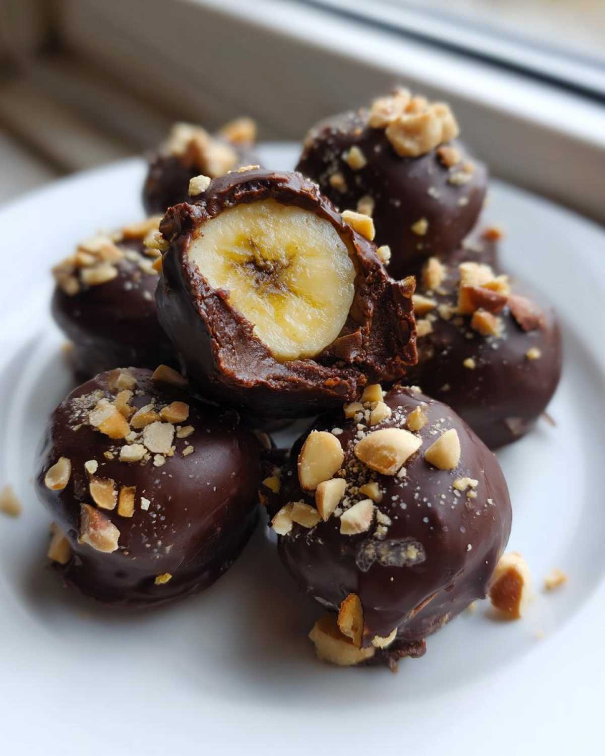A stack of No-Bake Chocolate Covered Banana Bites topped with chopped nuts, one is cut open showing the banana center.