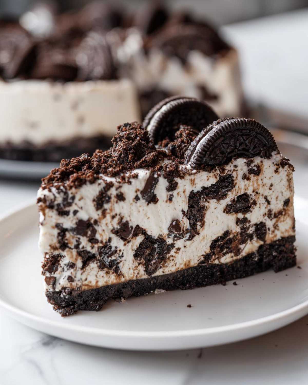 Close-up of a rich slice of No-Bake Oreo Cheesecake on a white plate, topped with crumbs and whole cookies.
