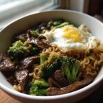 A close-up of One-Pot Beef and Broccoli Ramen Noodles topped with a sunny-side-up fried egg in a white bowl.