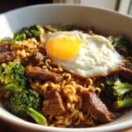 A close-up of One-Pot Beef and Broccoli Ramen Noodles topped with a sunny-side-up egg in a white bowl.
