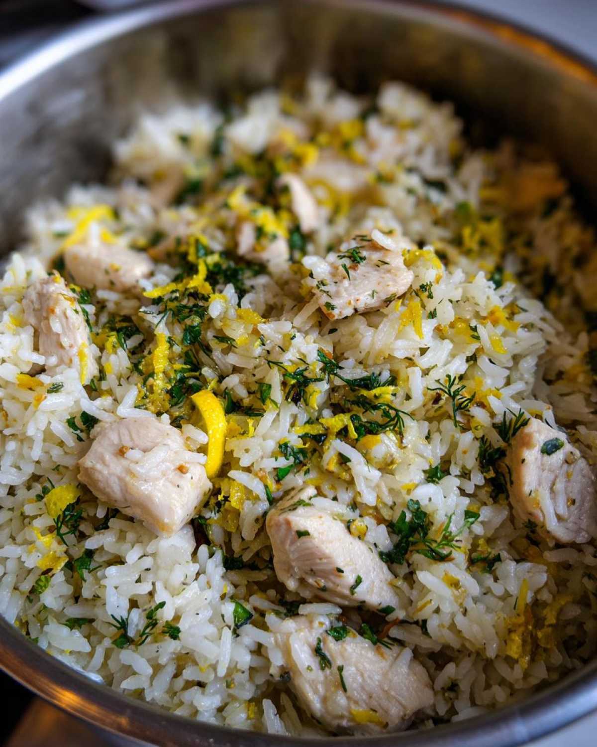 Close-up of fluffy white rice mixed with chunks of chicken, lemon zest, and fresh green herbs in a pot.