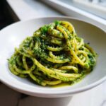 A close-up of thick pasta coated generously in bright green Vegan Basil Pesto, sprinkled with vegan parmesan.