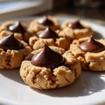 A close-up shot of several freshly baked Peanut Butter Blossom Cookies arranged on a white plate, each topped with a chocolate kiss.