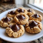 A close-up of several freshly baked Peanut Butter Blossom Cookies topped with melted chocolate kisses on a white plate.
