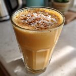 A tall glass of creamy, orange-hued Pineapple Coconut Smoothie topped with toasted coconut flakes and cinnamon.