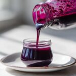 A glass jar is being filled with rich, dark purple Homemade Blueberry Syrup being poured from a larger bottle.