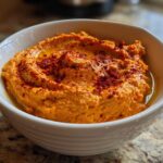 A close-up of vibrant orange Roasted Red Pepper Hummus topped with olive oil and paprika in a white bowl.