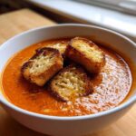 A white bowl filled with creamy Roasted Tomato Basil Soup, topped with three golden Grilled Cheese Croutons.
