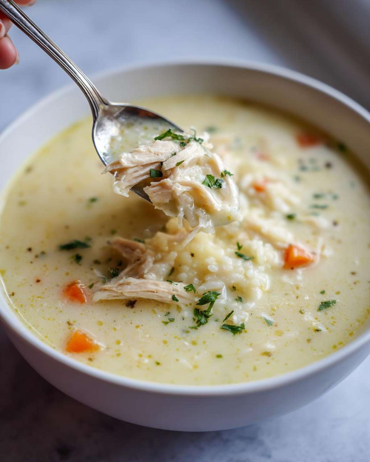 A spoonful of shredded chicken and rice being lifted from a bowl of creamy Greek Lemon Chicken Soup, garnished with parsley.