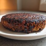 A single, firm Smoky Black Bean Burger Patty with a dark, crispy sear resting on a white plate.