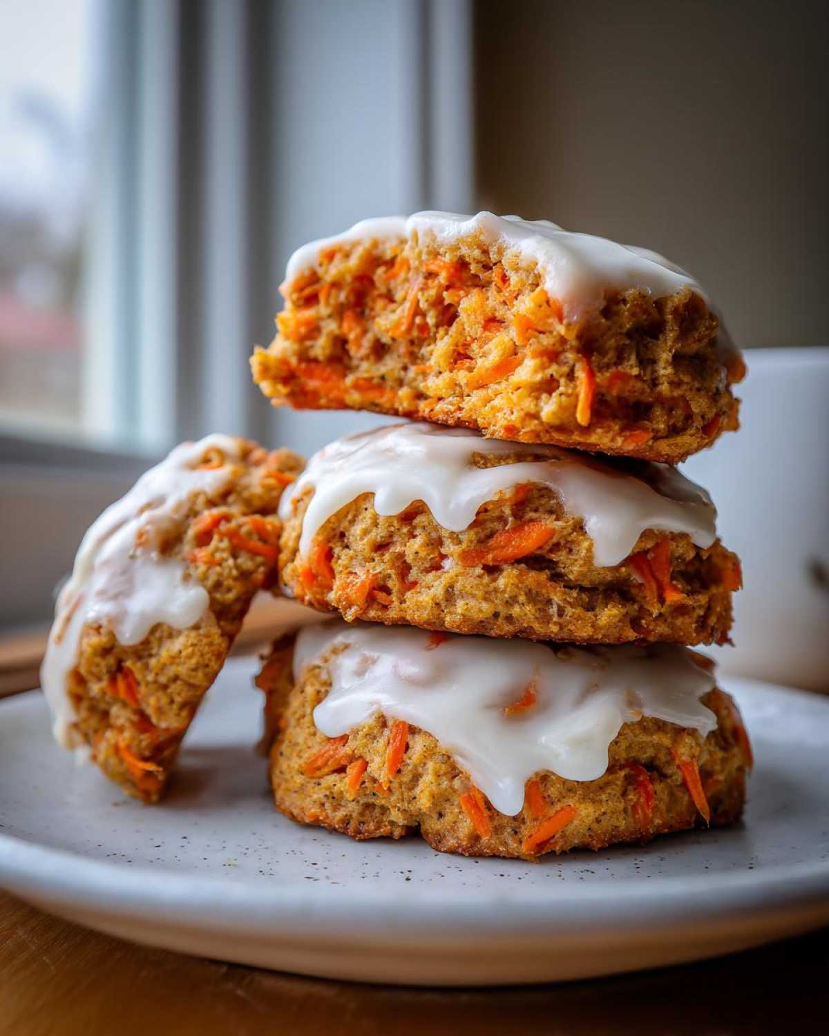 A stack of three Soft-Baked Carrot Cake Cookies topped with thick cream cheese glaze, one cookie is broken open.