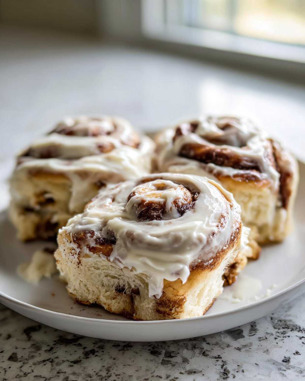 Three soft Gluten-Free Cinnamon Rolls piled high with thick, white cream cheese frosting on a white plate.