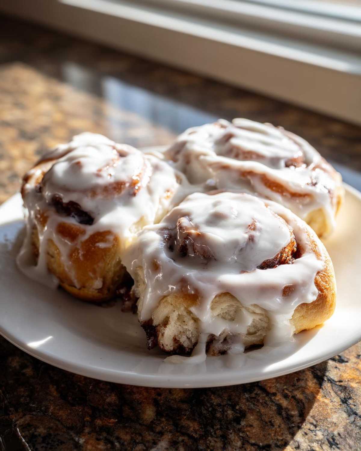 Three freshly baked Gluten-Free Cinnamon Rolls generously covered in thick white icing on a white plate.
