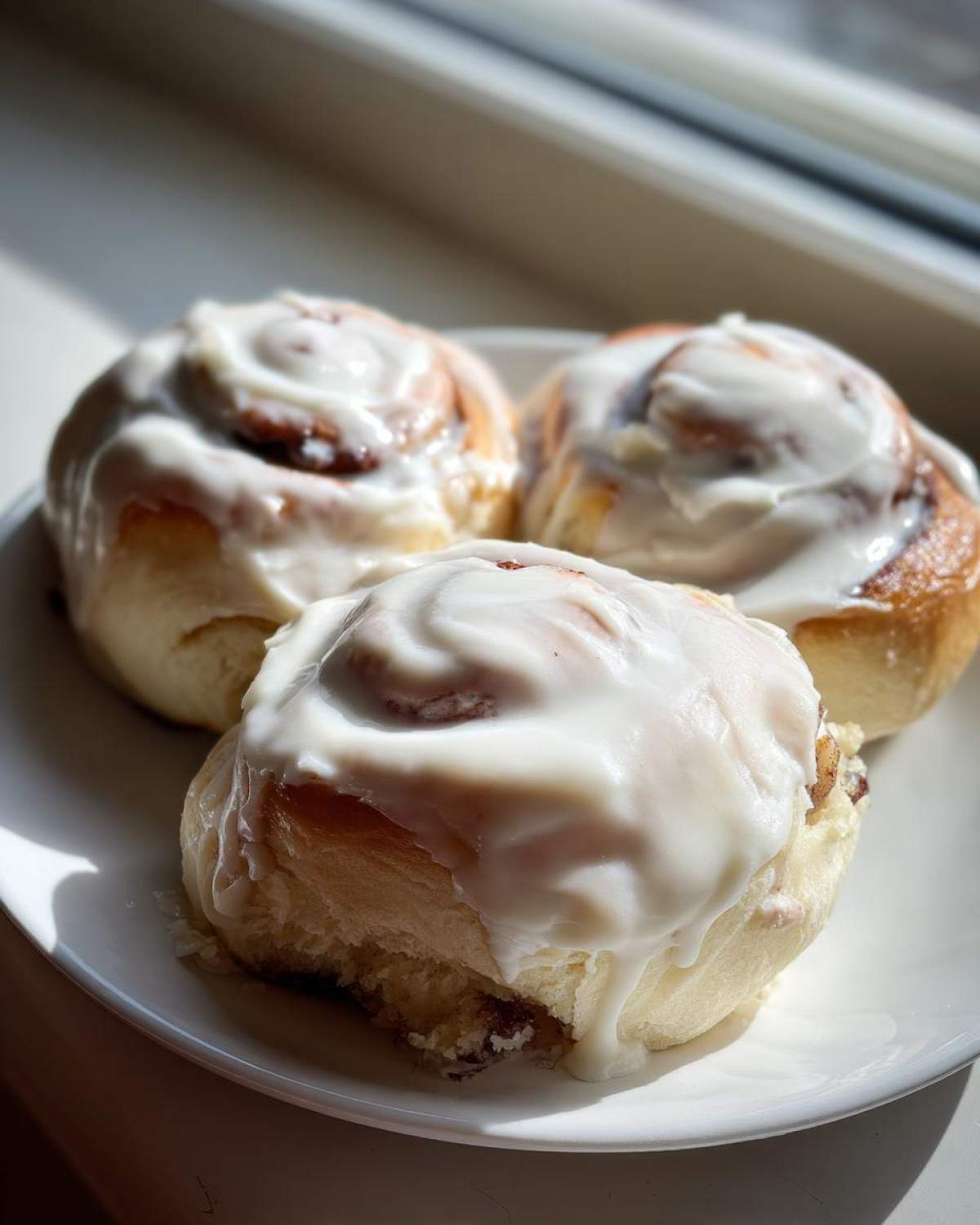 Three soft Gluten-Free Cinnamon Rolls covered in thick white icing, sitting on a white plate near a window.