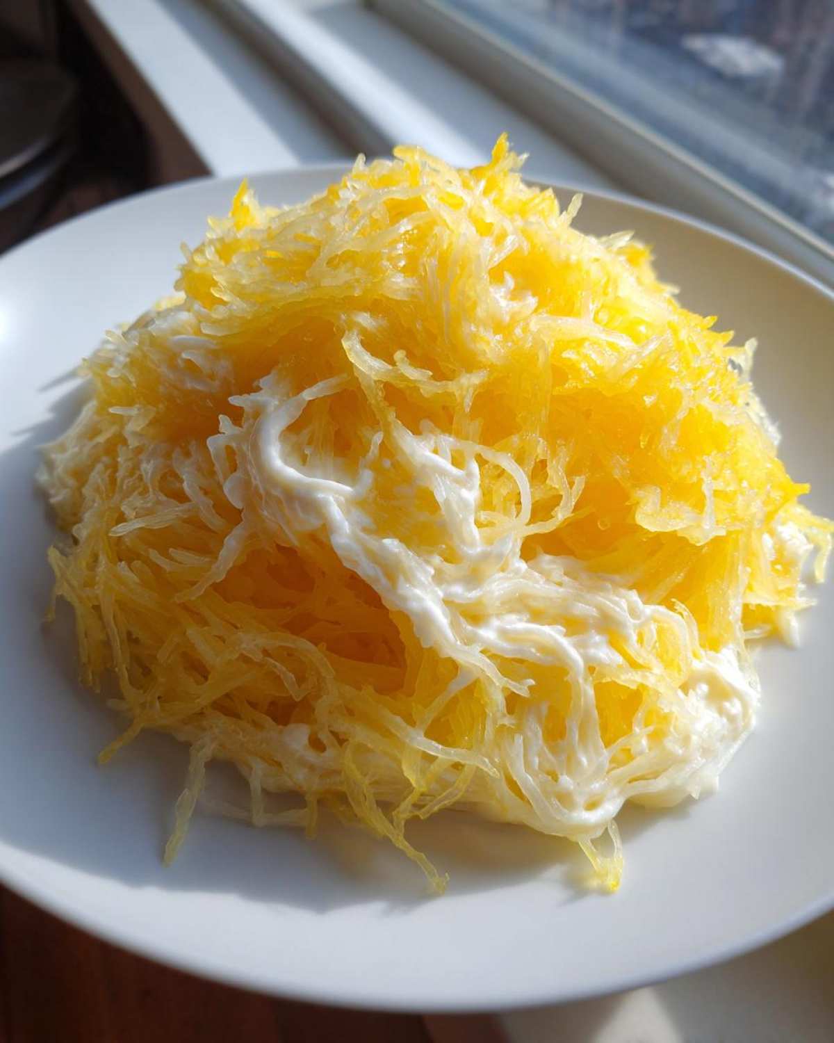 A close-up of a serving of bright yellow Spaghetti Squash Alfredo, tossed lightly with a creamy white sauce, served on a white plate.