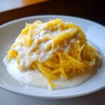 A mound of bright yellow spaghetti squash strands topped with creamy white Alfredo sauce, served in a white bowl.