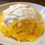 A close-up of bright yellow spaghetti squash strands topped with a generous dollop of creamy white Alfredo sauce in a white bowl.