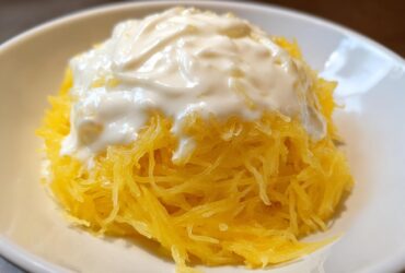 A close-up of bright yellow spaghetti squash strands topped with a generous dollop of creamy white Alfredo sauce in a white bowl.