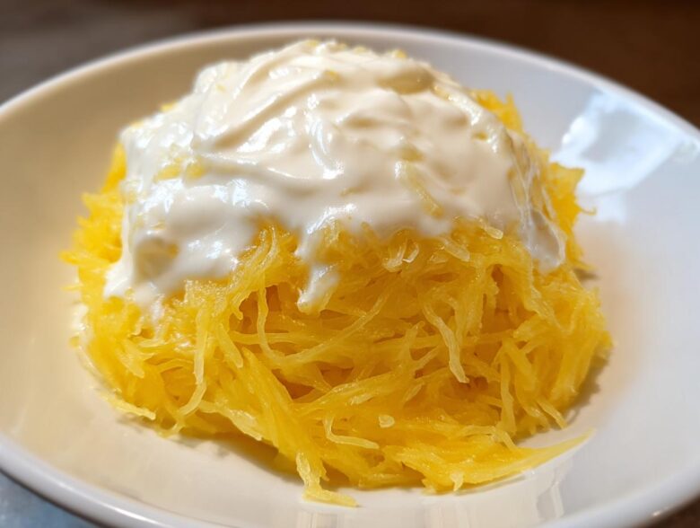 A close-up of bright yellow spaghetti squash strands topped with a generous dollop of creamy white Alfredo sauce in a white bowl.