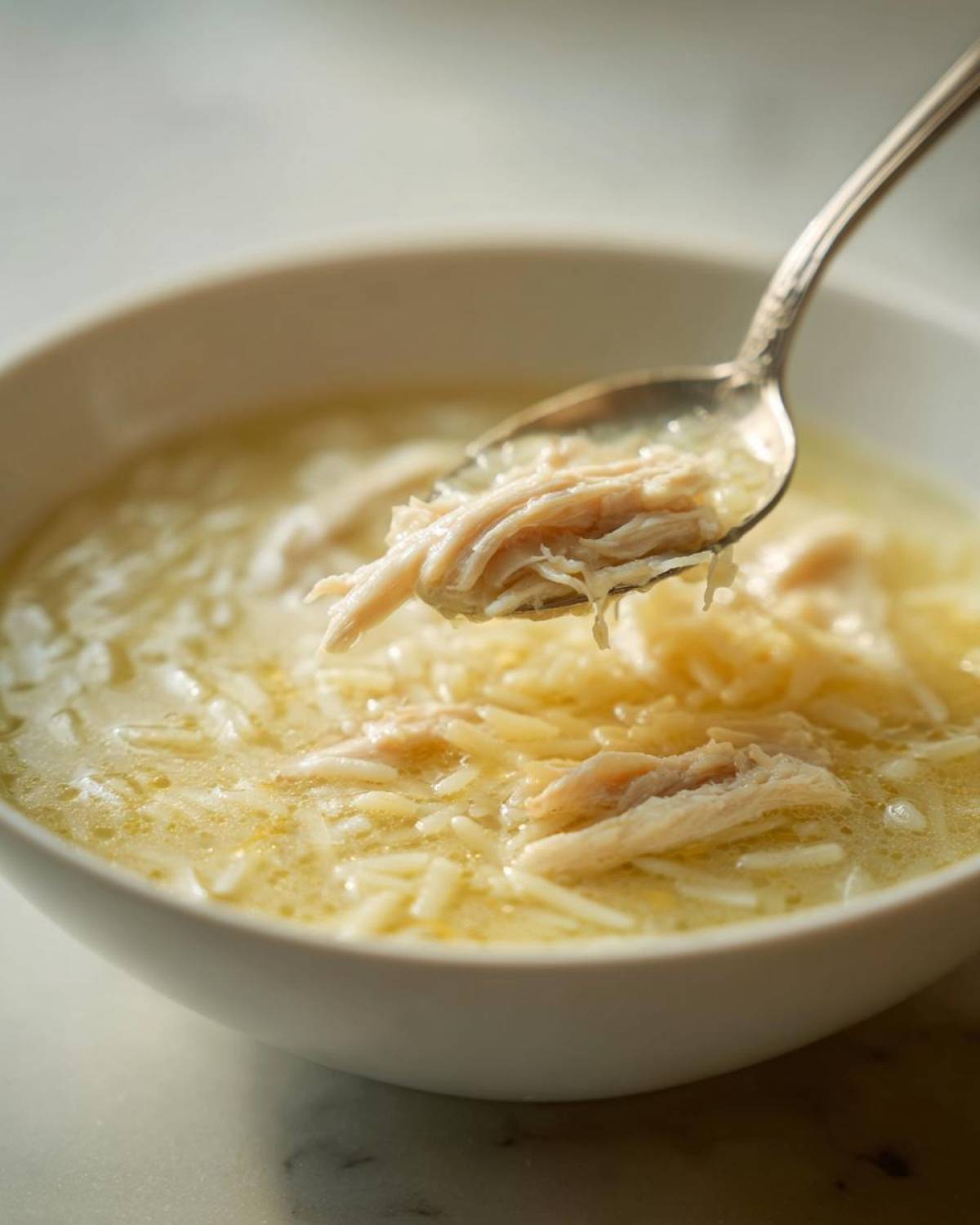 Close-up of a spoonful of shredded chicken being lifted from a bowl of creamy Greek Lemon Chicken Soup with orzo pasta.