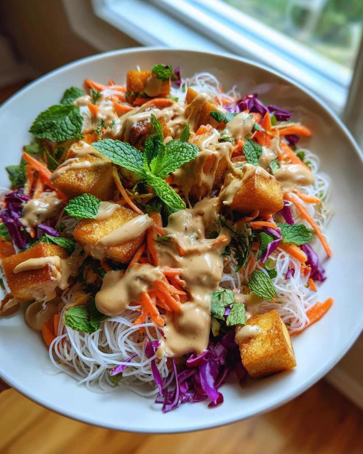 A close-up of a colorful Spring Roll Bowl with Peanut Sauce, featuring rice noodles, crispy tofu, shredded carrots, red cabbage, and fresh mint.