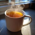Close-up of a steaming white mug filled with warm Honey Lemon Ginger Tea, backlit by sunlight.