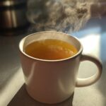A close-up of a steaming white mug filled with golden Honey Lemon Ginger Tea, highlighted by sunlight.