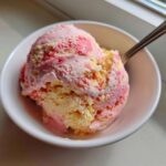 A close-up of a scoop of marbled Strawberry Banana Ice Cream in a white bowl with a spoon.