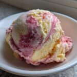 A close-up of two scoops of swirled Strawberry Banana Ice Cream, featuring yellow banana and pink strawberry swirls, served in a white bowl.