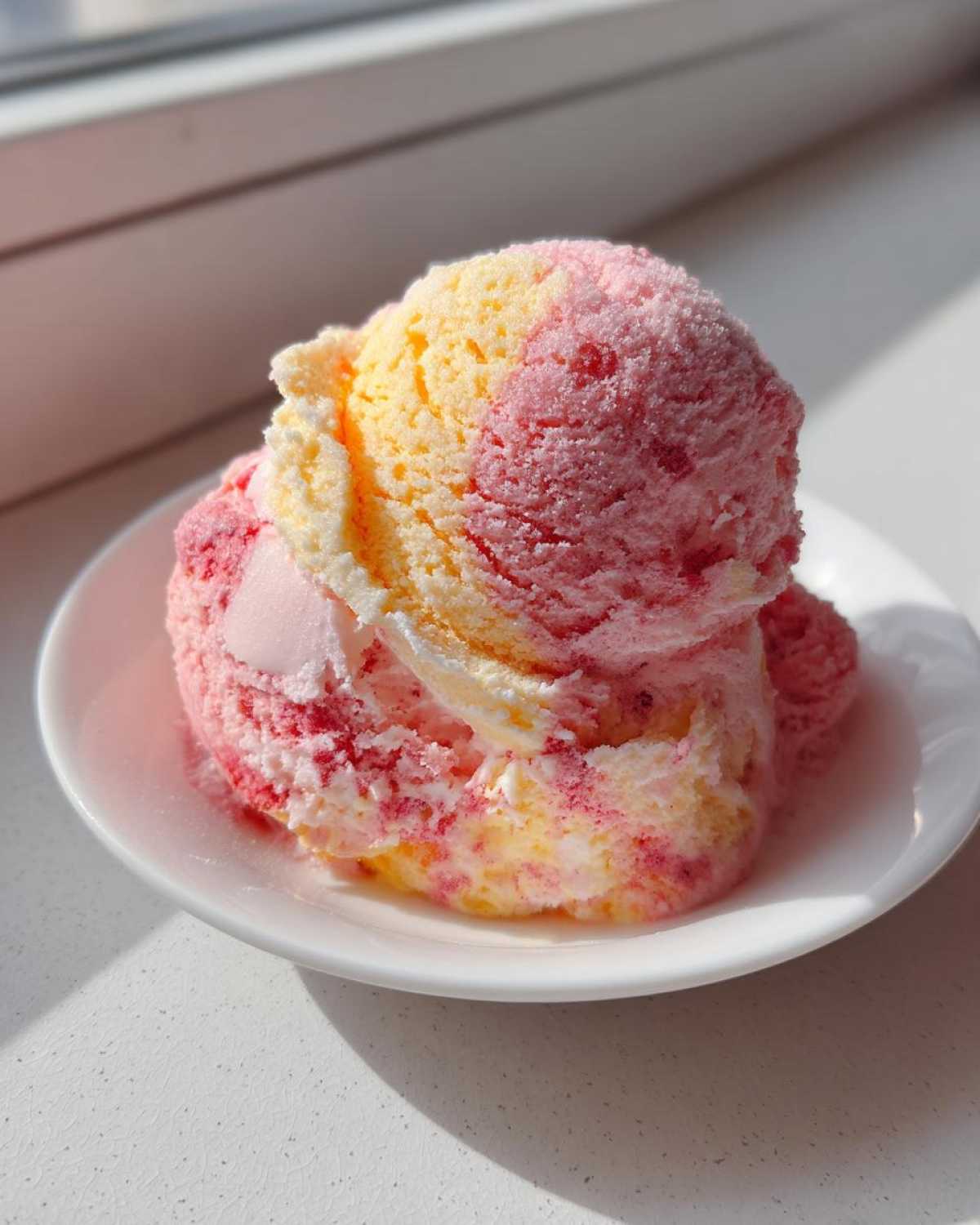 Two scoops of swirled pink and yellow Strawberry Banana Ice Cream served on a small white dish in bright sunlight.