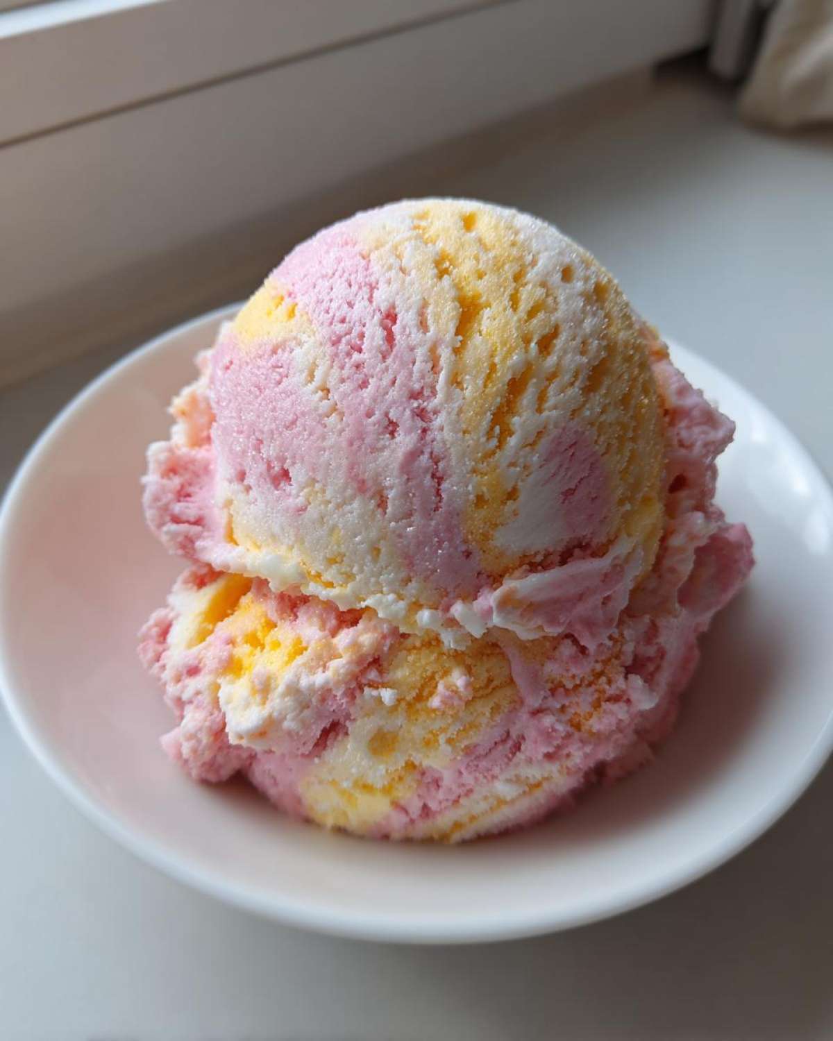 Two scoops of marbled pink and yellow Strawberry Banana Ice Cream served in a small white bowl.