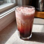 A tall glass filled with layered Strawberry Cream Cold Brew, showing dark coffee at the bottom and pink foam on top, sitting by a sunlit window.