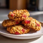 A stack of four golden Strawberry Oatmeal Breakfast Cookies studded with bright red strawberry pieces on a white plate.