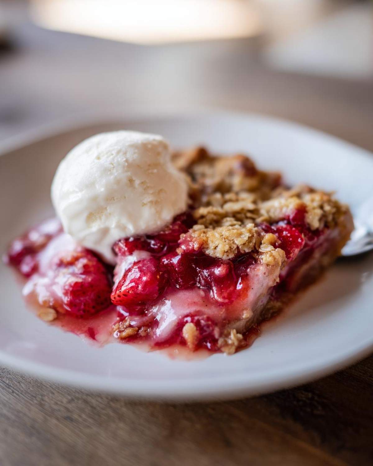 A warm slice of Strawberry Rhubarb Crisp topped with a scoop of melting vanilla ice cream on a white plate.