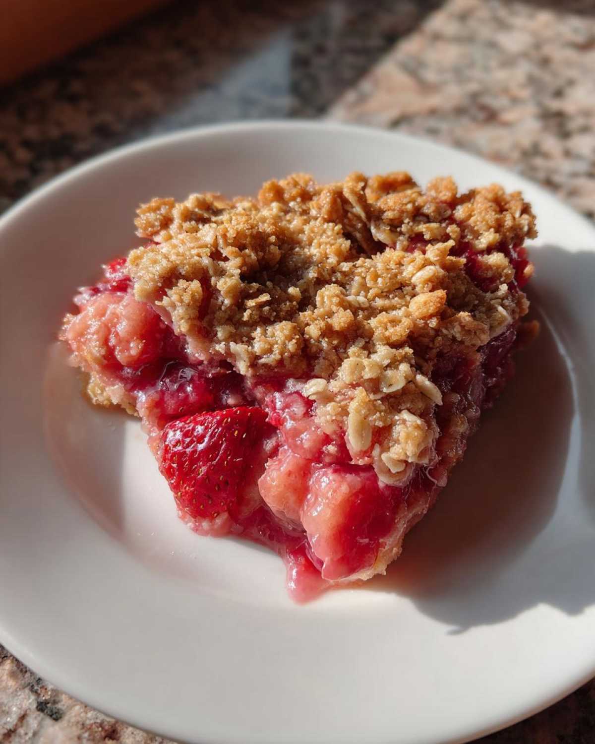 A close-up of a warm slice of Strawberry Rhubarb Crisp with a golden oat topping on a white plate.
