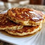 A close-up of three golden-brown Vegan Banana Oat Pancakes stacked and drizzled with syrup on a white plate.