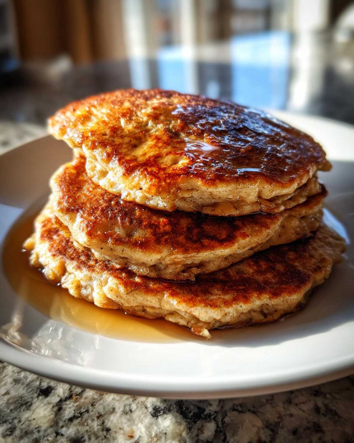 A close-up stack of four golden brown Vegan Banana Oat Pancakes drizzled with syrup.