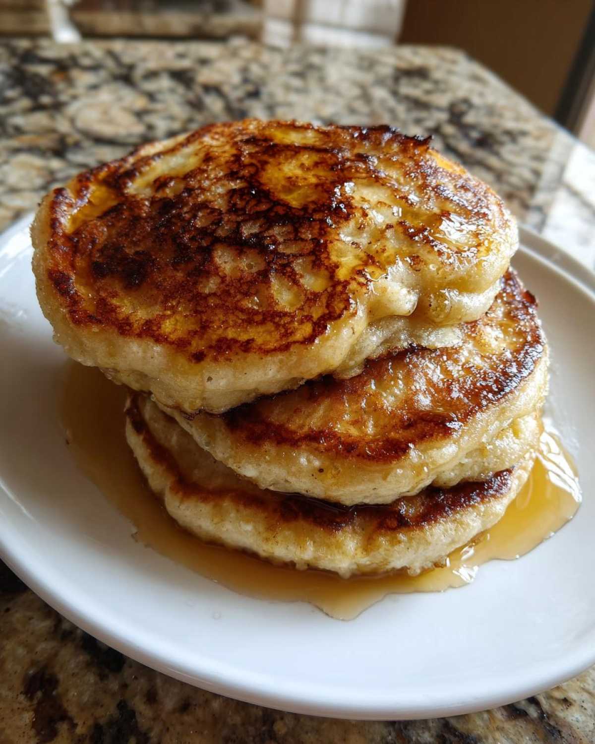 Close-up of a stack of three golden-brown Vegan Banana Oat Pancakes drizzled with syrup on a white plate.