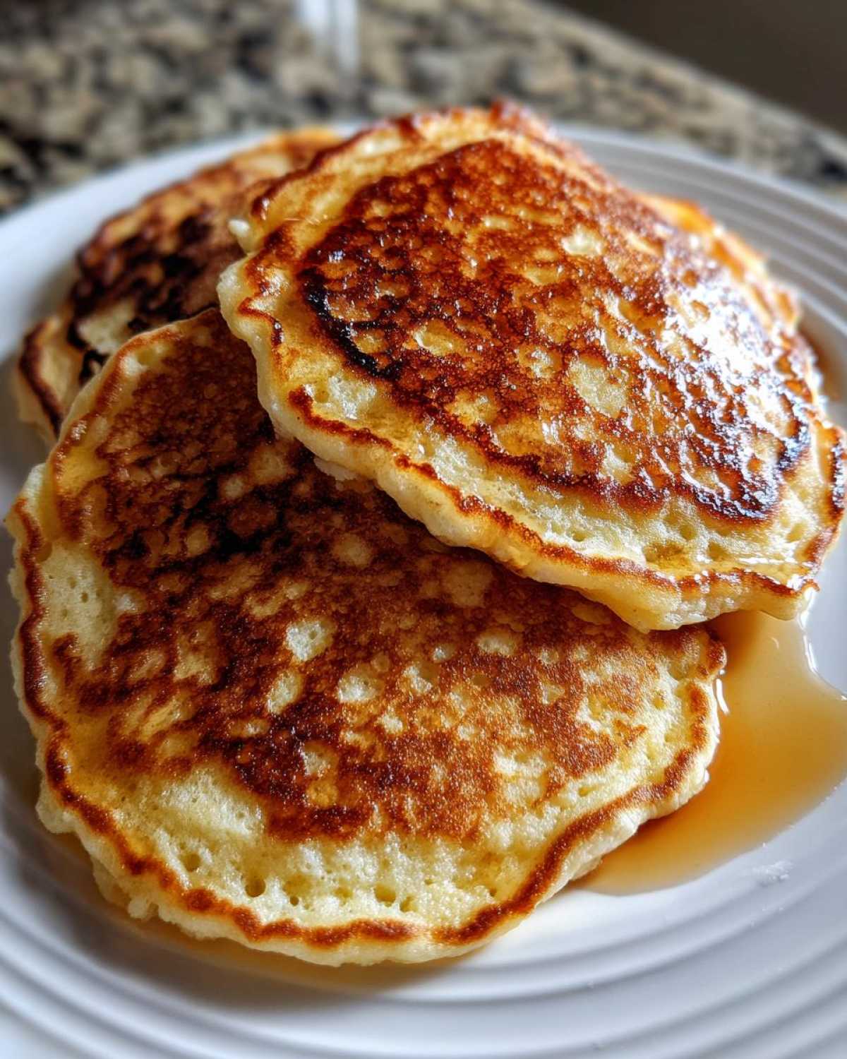 Close-up of a stack of golden-brown Vegan Banana Oat Pancakes drizzled with syrup on a white plate.