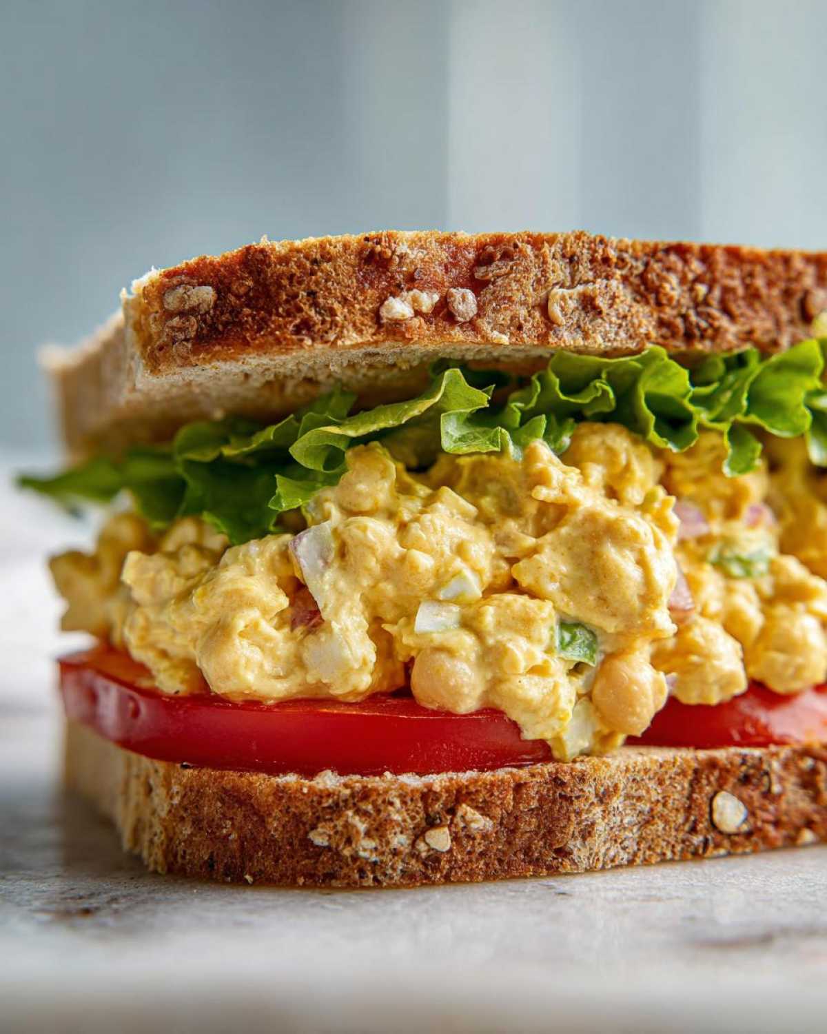 A close-up of a delicious Vegan Chickpea Salad Sandwich layered with creamy filling, tomato, and lettuce on whole grain bread.