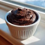 A close-up of rich, dark Vegan Chocolate Avocado Mousse served in a small white fluted ramekin near a window.